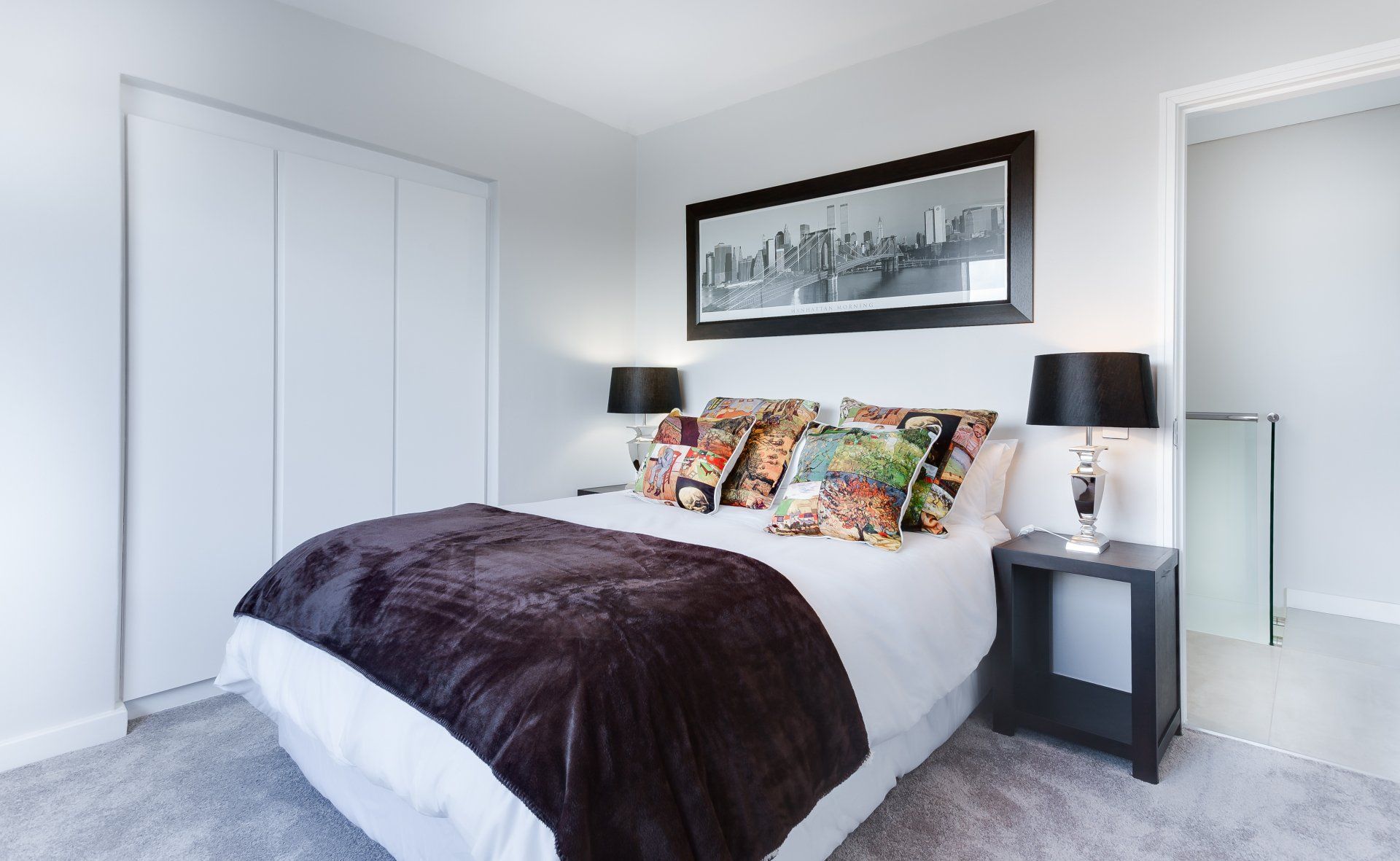 Bright bedroom with a neatly made bed, dark blanket, colorful pillows, and black bedside lamps