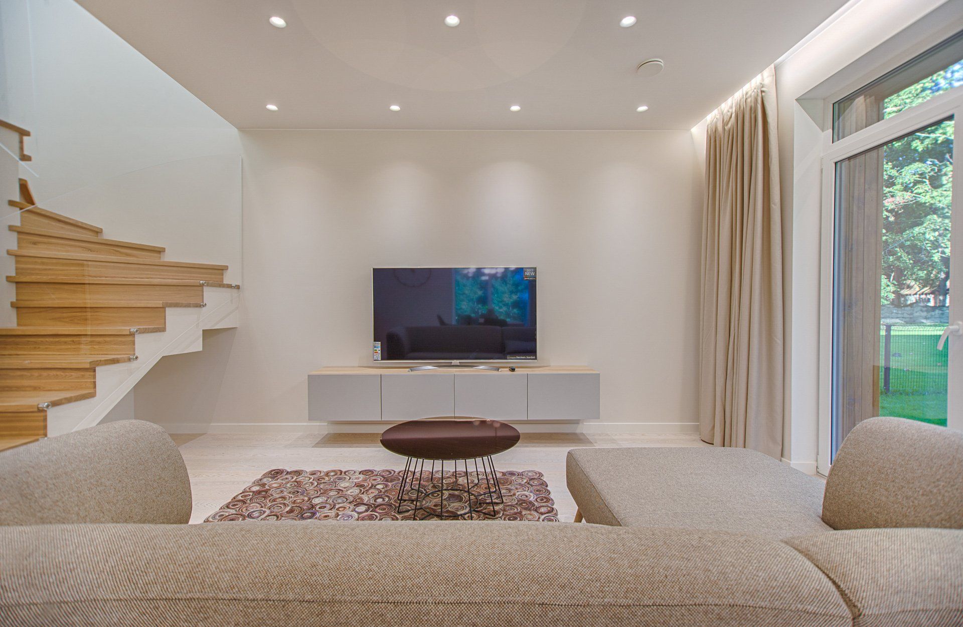Modern living room with a sectional sofa, wall-mounted TV, floating stairs, and large windows overlooking greenery